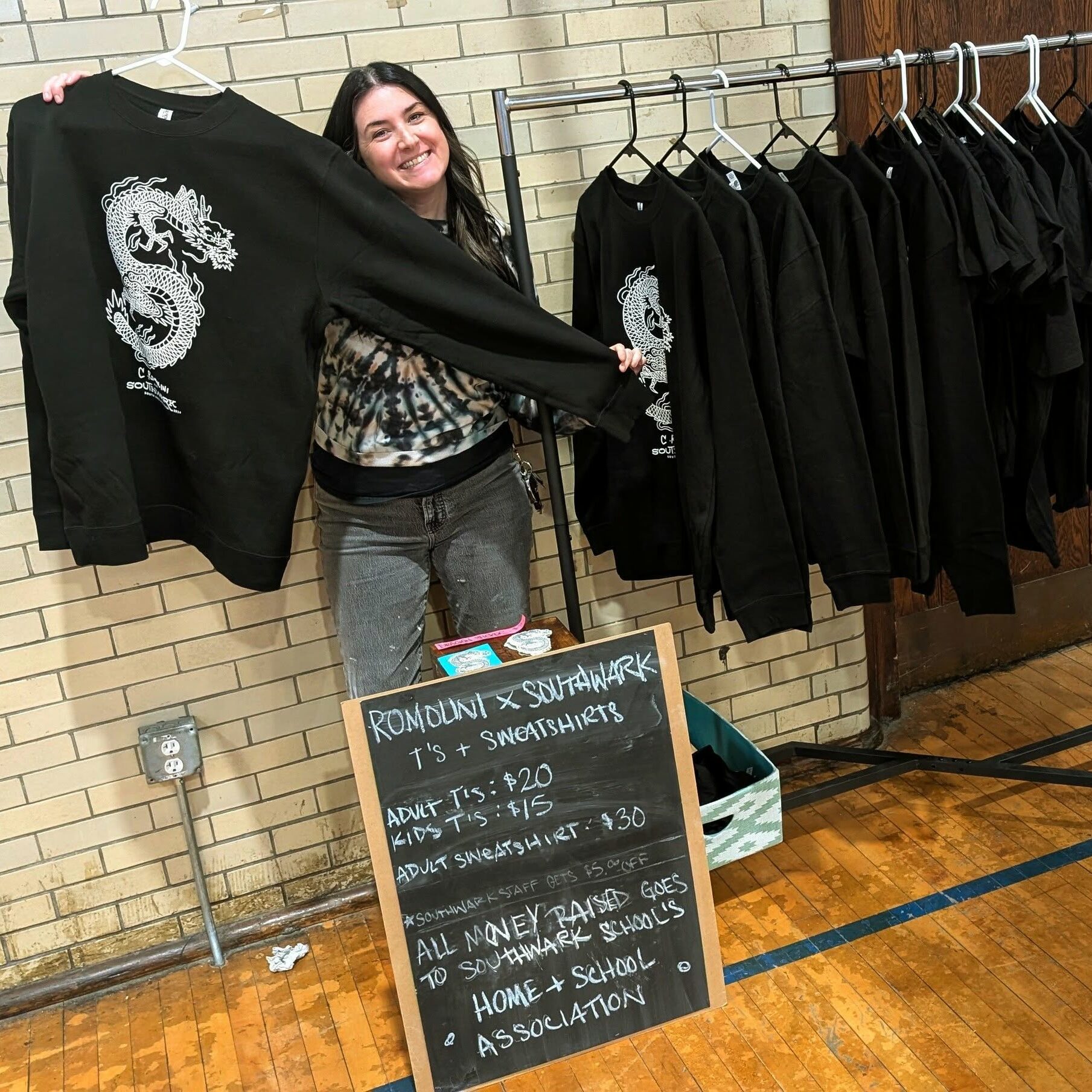 A Southwark School Home & School Association volunteer displays fundraiser sweatshirts, with proceeds supporting school programs and student activities.